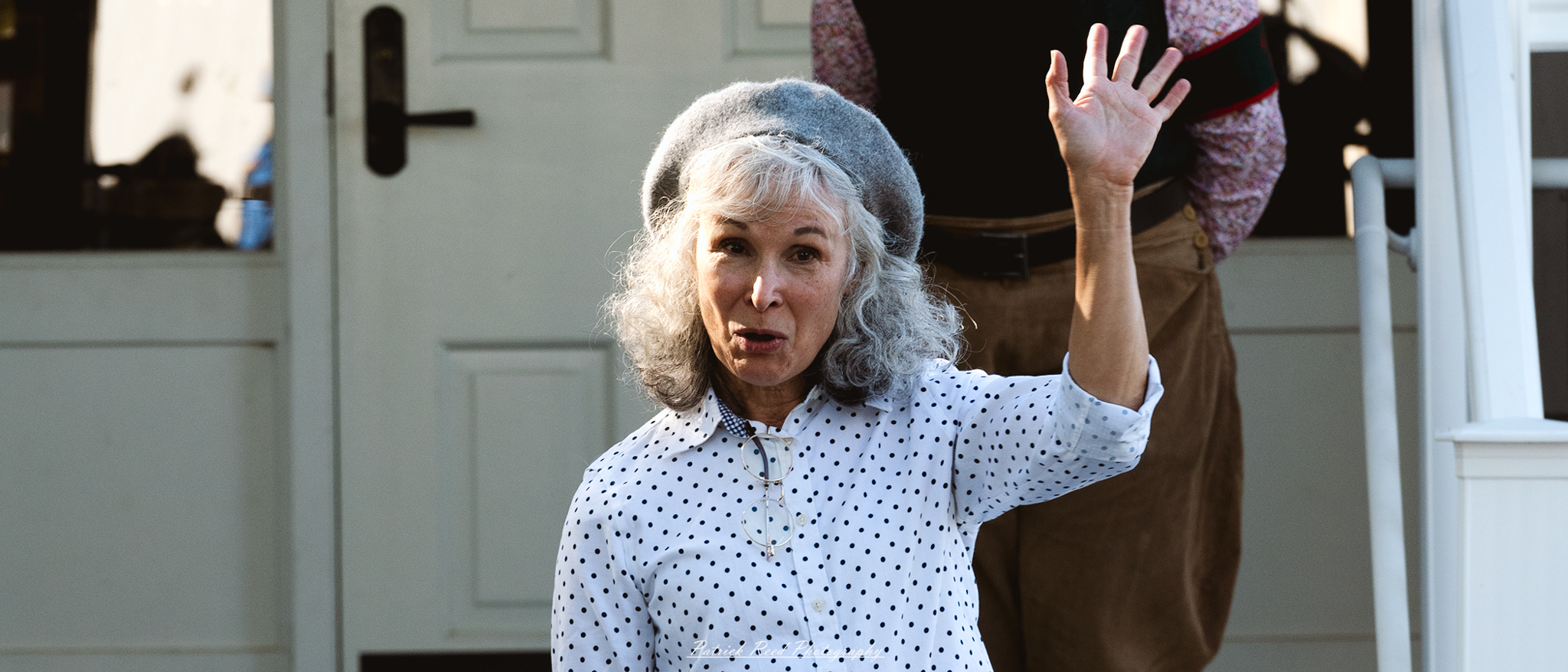 An old woman dressed in 1940s clothing stands with a warm smile, waving at the camera. Her attire includes a patterned dress with a cinched waist, a modest cardigan, and a vintage hat adorned with flowers. The background features a quaint, sunlit street or garden, adding a nostalgic charm to the scene. Her kind expression reflects the friendliness of the era, evoking feelings of warmth and community.